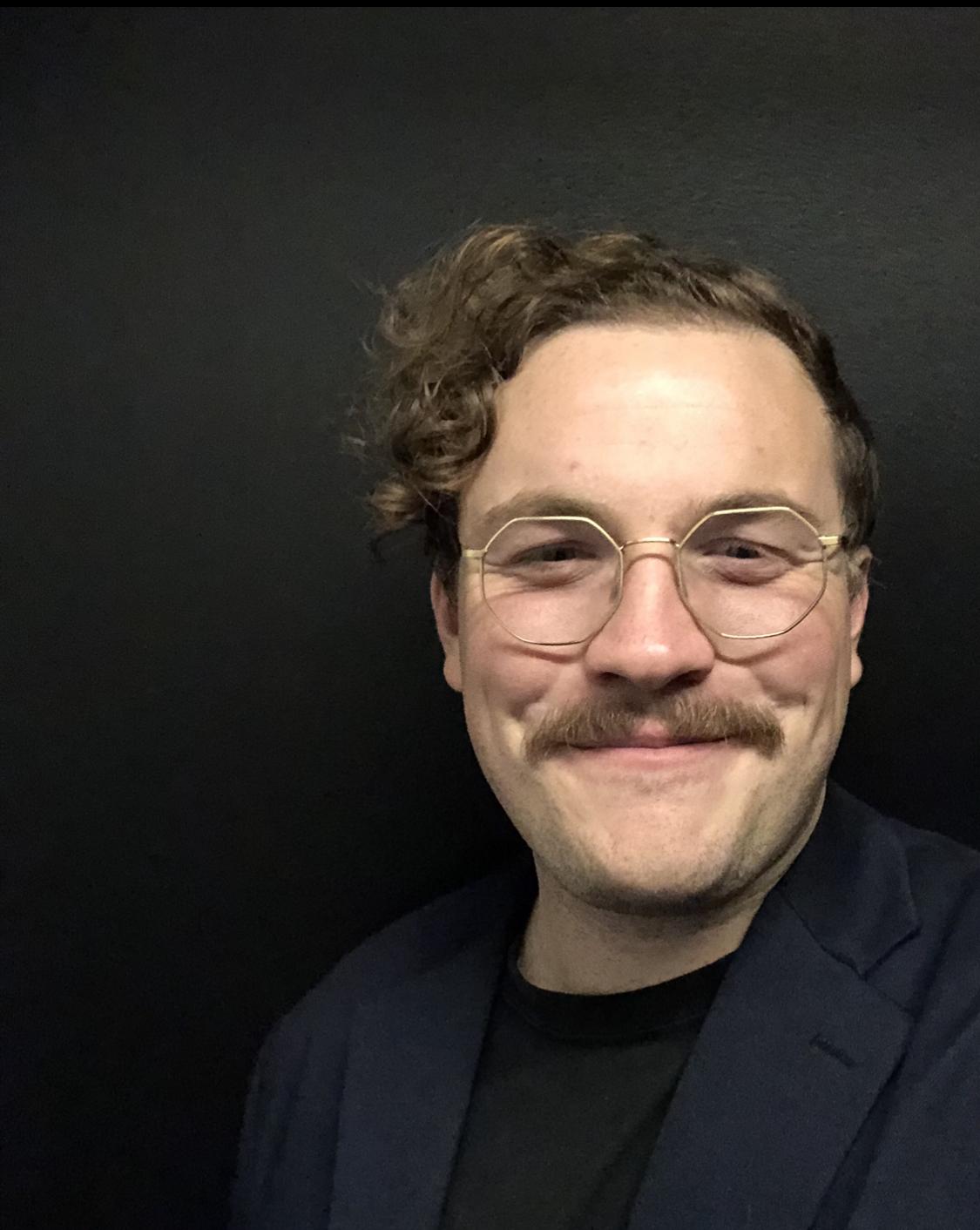 Zac Taylor pictured in the fore, wearing glasses and with a moustache. Black background. 