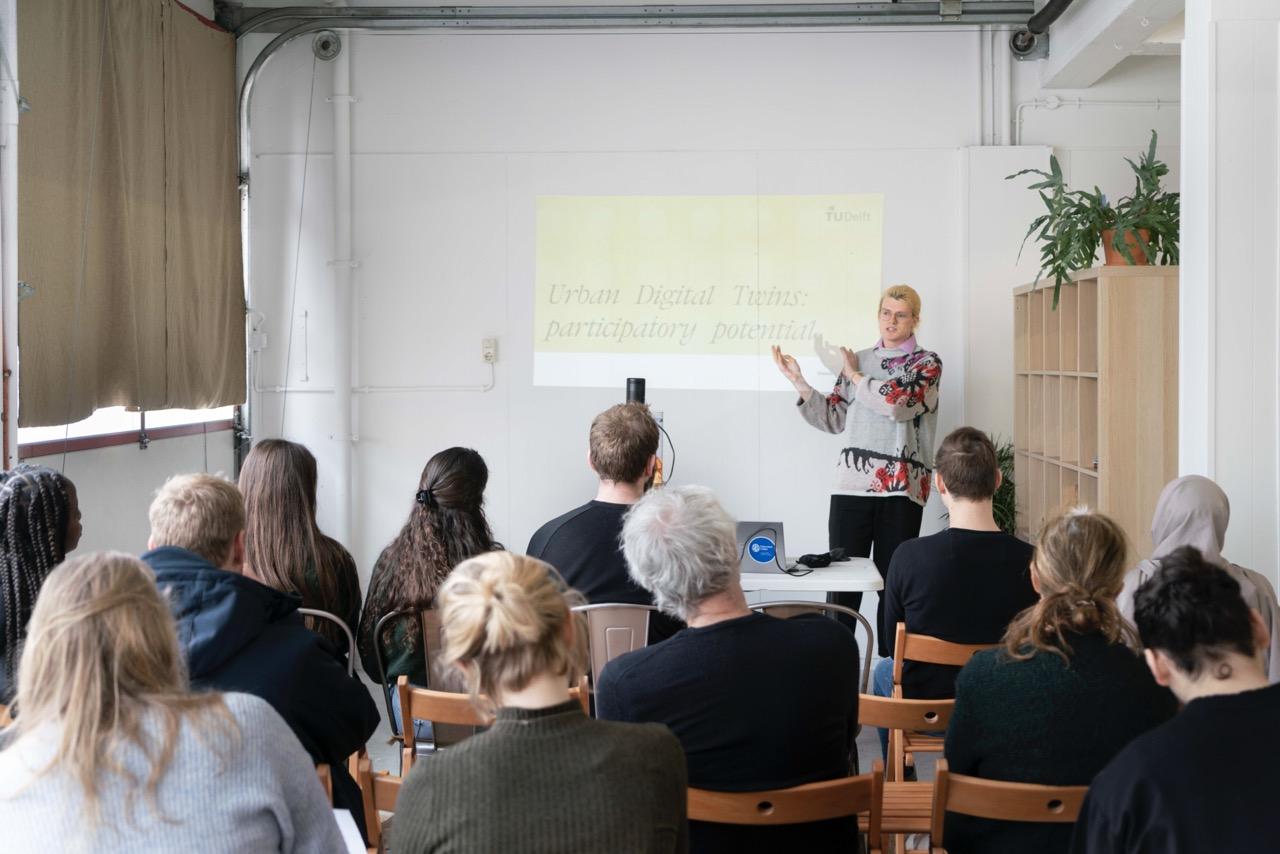 David delivers their presentation to students, dressed in a floral sweater. Audience is in the fore of the image