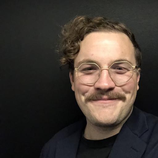 Zac Taylor pictured in the fore, wearing glasses and with a moustache. Black background. 
